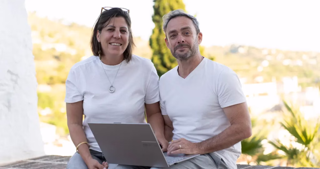 A candid photograph of Mik and Fefe smiling and working together on laptops in their home office, celebrating one year of business.